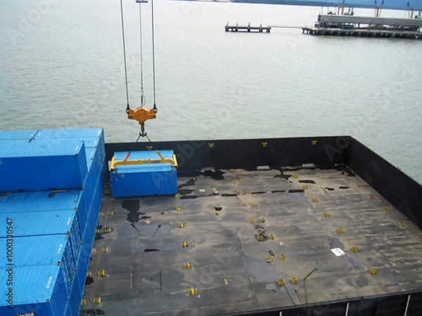 Obraz A heavy-duty industrial crane lifting a large blue shipping container at a busy port, set against a backdrop of a bright blue sky and ocean. The scene highlights the efficient cargo handling process