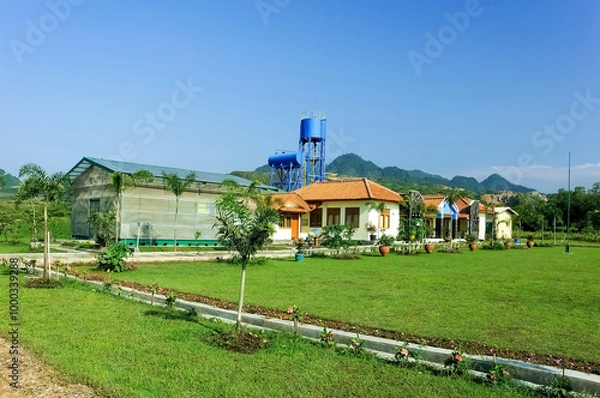 Obraz A serene villa surrounded by lush greenery, featuring a traditional yet modern architectural design with a sloping red-tiled roof. The villa is nestled amidst a garden landscape