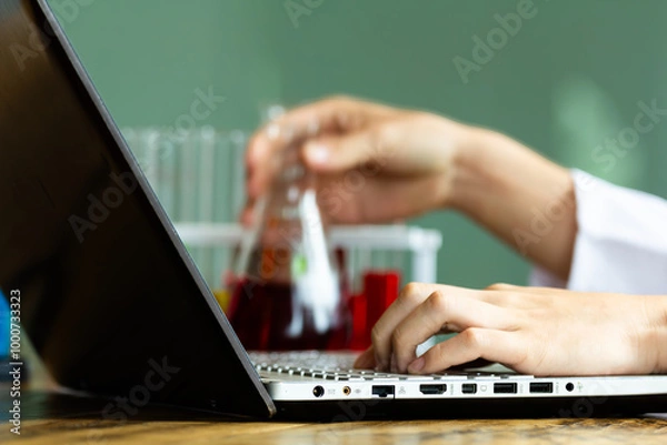 Obraz Close up male lab assistant chemist using laptop for data entry and in other hand holding test tube with red blood sample while sitting in central laboratory.