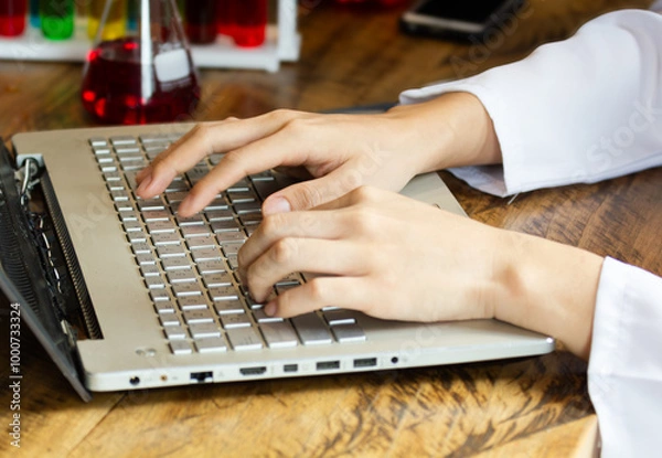 Fototapeta Close up male lab assistant chemist using laptop for data entry and in other hand holding test tube with red blood sample while sitting in central laboratory.