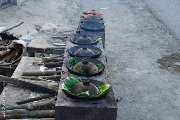 Obraz A traditional Indonesian serabi-making process, showing hands pouring batter onto a clay pan over a wood-fired stove. The smoky atmosphere and authentic tools emphasize the local street food culture.