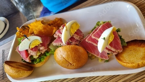 Fototapeta A gourmet dish featuring three small sandwiches topped with seared tuna, lettuce, a slice of hard-boiled egg, and a sprig of herb, served with crispy golden-brown chips on a white rectangular plate.