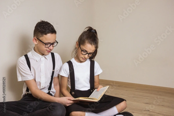 Fototapeta A boy and a girl in a school uniform and glasses sit on the floor and read a book