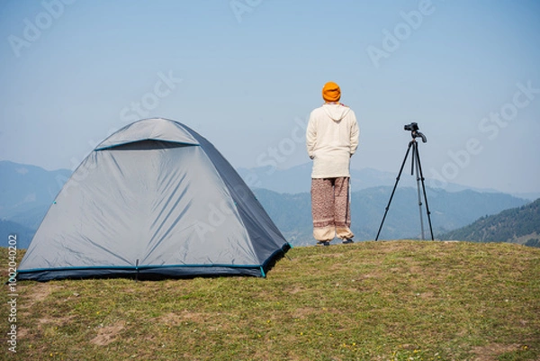 Fototapeta An adventurer stands beside a camping tent, looking at the mountain view — a perfect moment of travel and adventure.