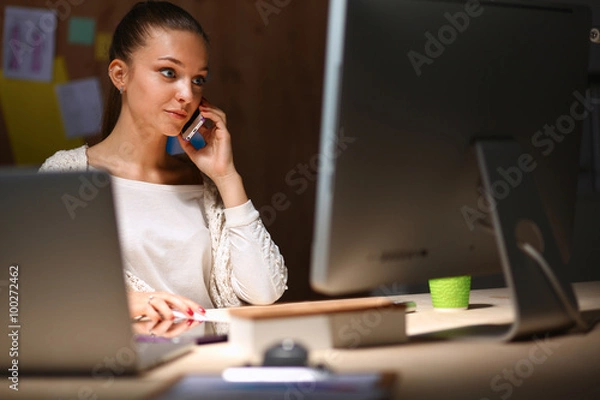 Obraz Young woman working in office, sitting at desk and talk on the phone