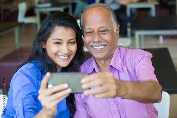 Obraz Closeup portrait happy elderly gentleman in pink shirt and lady in blue top taking selfie together, isolated indoors background. Say cheese and smile
