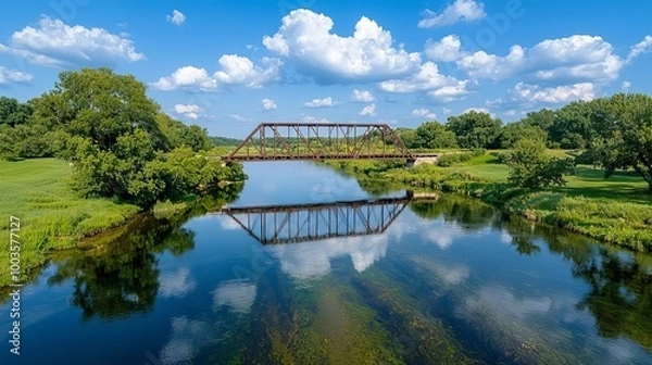 Fototapeta Railroad bridge over tranquil river reflecting blue sky and lush greenery.