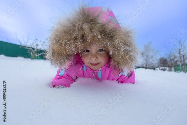 Fototapeta A child in a colorful jumpsuit lies in the snow and looks at the camera, smiling. The child is in the snow, wearing a hood with fluffy fur. The child is crawling in the snow, enjoying the winter.