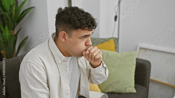 Fototapeta A young man coughing indoors while sitting on a couch, with a modern living room setting in the background.