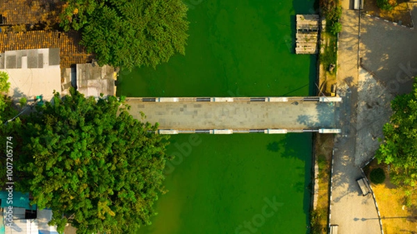 Fototapeta An aerial view captures the serene beauty of a footbridge over tranquil green waters, bordered by lush trees and urban structures