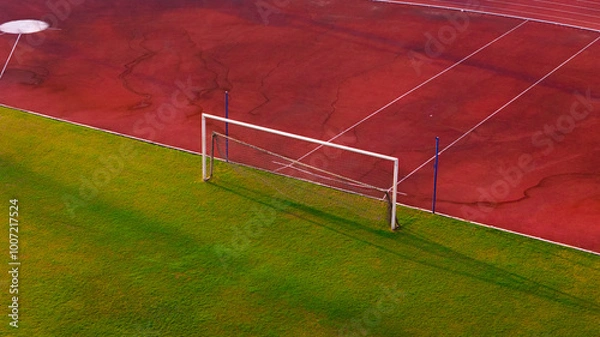Fototapeta A dynamic aerial view of a soccer goal post on a lush green field adjacent to a vibrant red running track, capturing the essence of outdoor sports and athletics
