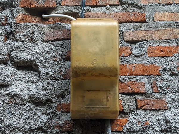 Obraz An old, weathered electrical box mounted on a rough, textured brick wall. The contrast between the worn yellow box and the rustic wall creates an industrial and urban aesthetic.