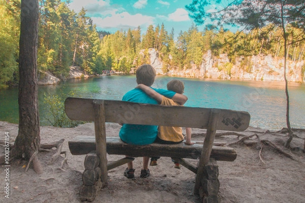 Obraz Two brothers sitting on wooden deck chair in the rays of summer sun near lake in rocks Adrspach in the Czech republic. Blue sky, forest and lake. Summer concept.