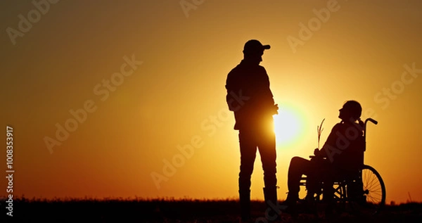 Fototapeta Silhouette of a man assisting a person in a wheelchair against a vibrant sunset backdrop.