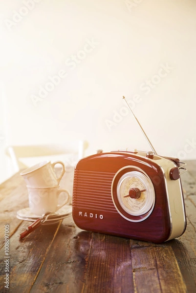 Obraz A retro-style radio is on a wooden table next to porcelain white mugs, illuminated by warm sunlight. Morning tea. Vertical photo.