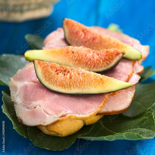 Fototapeta Fresh fig slices on smoked ham sandwich on fig leaves on blue wood (Selective Focus, Focus on the first fig slice)