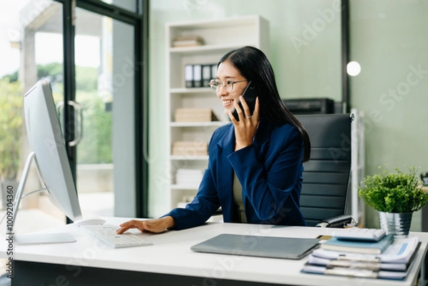 Fototapeta Asian woman sitting at a desk using a laptop computer Navigating Finance and Marketing