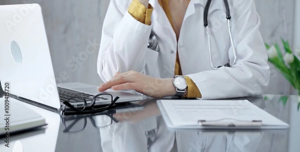 Fototapeta Thinking doctor woman is reviewing medical records on laptop. Close-up of a healthcare unknown professional analyzing patient information on a computer. Medicine