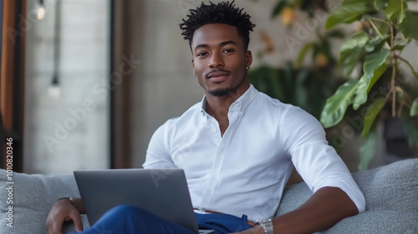 Fototapeta African-American man wearing a white shirt and jeans, sitting on a grey sofa and using a laptop indoors. She is looking at the camera and smiling, showcasing a home interior design concept