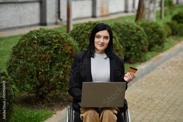 Fototapeta A woman in a wheelchair uses a laptop and a credit card while sitting on a path in a park.