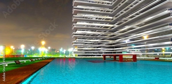 Obraz Waterfront with benches and lawn lights along a large azure pool on the grounds of an elite home against the backdrop of a nighttime village. 3D rendering.