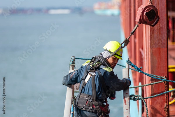 Fototapeta A deckhand prepares the pilot ladder as the ship readies for port entry. Secured in a safety harness, he carefully ensures the setup is ready, focusing on the task in a high-risk environment. 