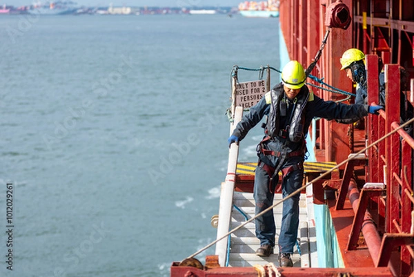 Fototapeta A deckhand prepares the pilot ladder as the ship readies for port entry. Secured in a safety harness, he carefully ensures the setup is ready, focusing on the task in a high-risk environment. 