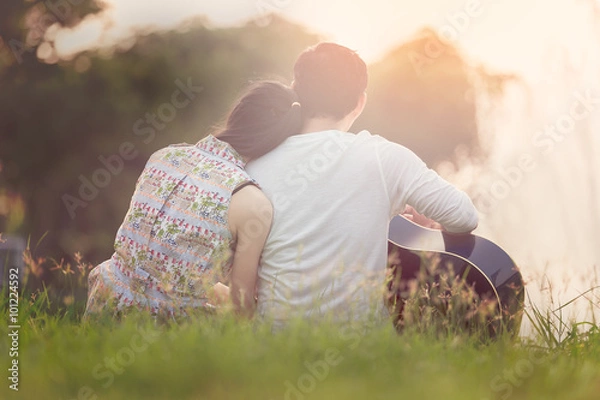 Obraz Lovers in a lake. Young couple in love sitting on the park ground near the water while these young man playing guitar in sunset time. Post processing in vintage style