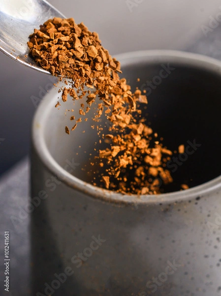 Obraz Grains of freeze dried instant coffee,falling from a silver tea spoon,into an empty cup,on a slate grey background..
