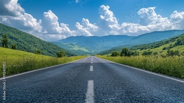 Fototapeta An asphalt road stretching towards green mountains, with a bright blue sky and puffy clouds on a sunny, clear day.