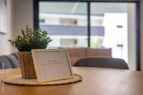 Fototapeta A framed welcome sign reading "Bienvenido a nuestra casa de verano" with a potted plant on a table in a modern summer home