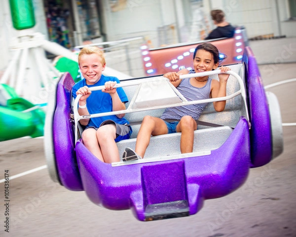 Fototapeta Two diverse kids riding an amusement park ride at the fun theme park. Screaming, laughing and having a great time on a thrilling carnival ride. Intentional motion blur on the image