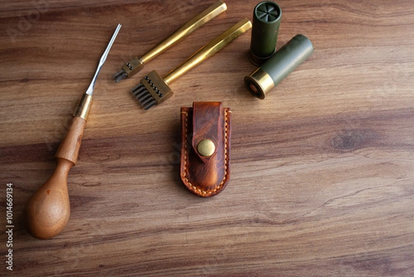 Fototapeta a handcrafted leather knife sheath with a brass button clasp, various leatherworking tools, all arranged on a polished wooden surface that highlights the artisanal and rugged aesthetic of the items