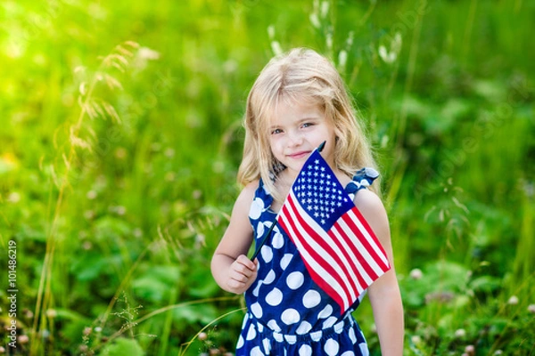 Obraz Cute smiling little girl with long curly blond hair holding an american flag on sunny day in summer park. Independence Day, Flag Day concept