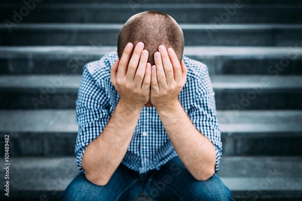 Fototapeta Outdoor portrait of sad young man covering his face with hands sitting on stairs. Selective focus on hands. Sadness, despair, tragedy concept