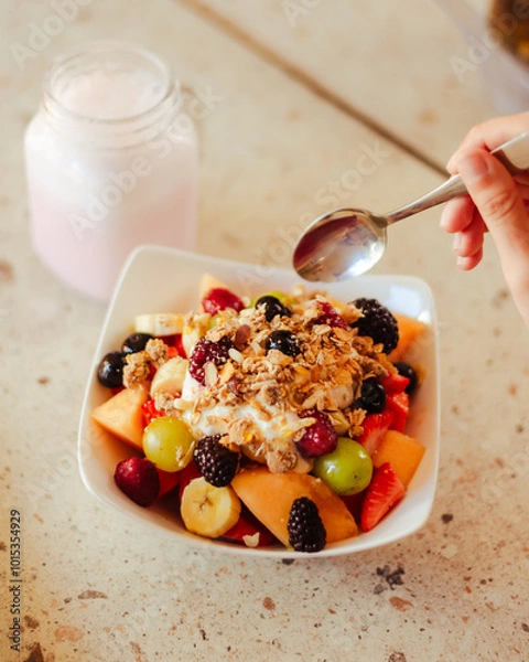 Fototapeta Mano sosteniendo una cuchara, debajo un bowl con frutos rojos y cereales de un desayuno saludable