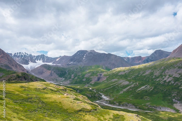 Fototapeta Wide green alpine valley with snake river among hills and rocks with vast view to large snow mountain top and big glacier tongue under cloudy sky. Serpentine river in high mountains. Shadows of clouds
