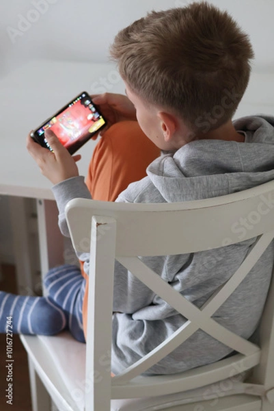 Obraz Teenage boy sitting on a chair at home and playing a game on his smartphone