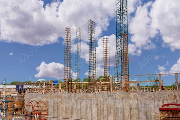 Fototapeta Construction site under a clear blue sky with clouds. Several tall, vertical steel rebar structures are in place for future concrete columns. In the foreground, concrete foundation.