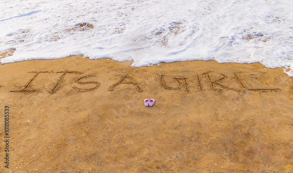 Fototapeta It's a boy and it's a girl baby written text announcement on the beach sand with shoe. Clean sea water and small beach waves are in background