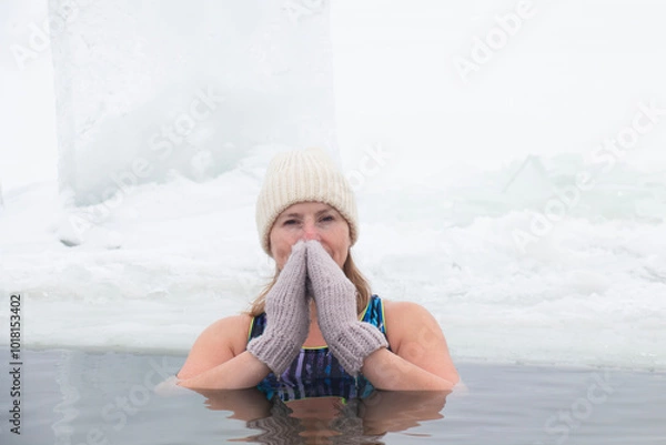Fototapeta A 50-year-old woman swims in a lake wearing a hat and mittens on a snowy, frosty, winter day, hardening and water treatments in an ice hole. A healthy lifestyle.