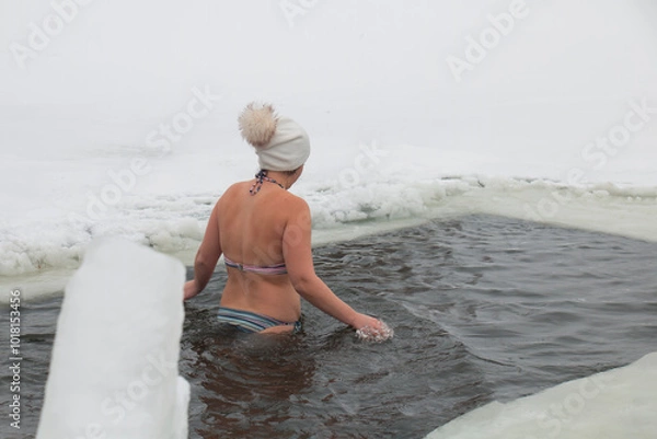 Fototapeta A woman swims in a lake in a hat, on a snowy, frosty, winter day, hardening and water procedures in an ice hole. Healthy lifestyle.