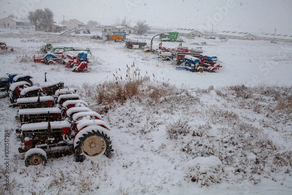 Fototapeta a winter scene on a farm where tractors and various colorful farm machines are covered in a layer of snow, emphasizing the stillness and inactivity brought by the snowy weather