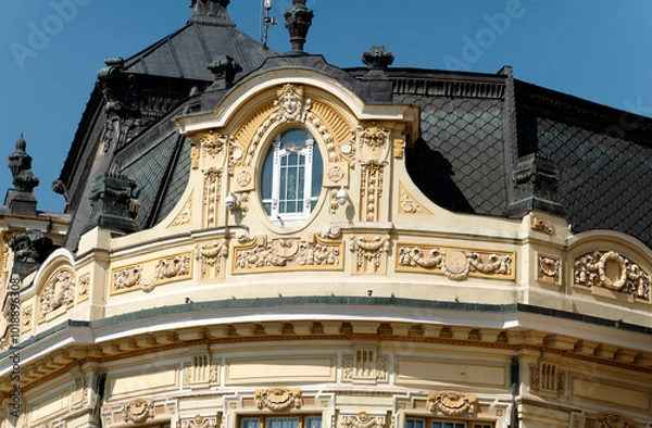 Fototapeta A close-up of a historic building facade featuring a curved window surrounded by intricate decorative elements. The building has a dark roof and a light-colored exterior.