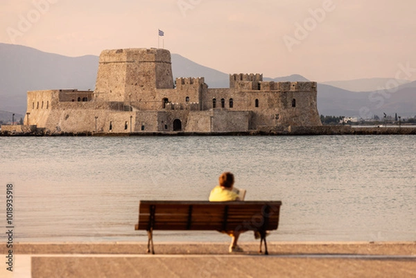 Obraz A solitary figure sits on a bench, gazing at the historic Bourtzi fortress in Nafplio, Greece, as the sun sets behind the serene waters, creating a tranquil atmosphere.