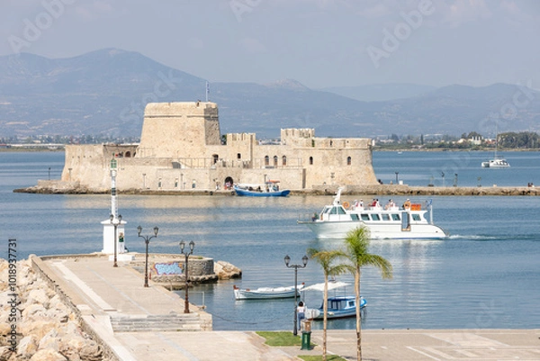 Obraz Bourtzi fortress stands majestically in the serene waters of Nafplio, surrounded by mountains. Local boats sail by, creating a picturesque coastal landscape that captures timeless beauty.