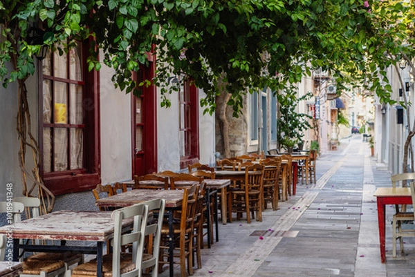 Obraz A serene street in Nafplio features rustic wooden tables lined under a canopy of lush vines, inviting guests to enjoy outdoor dining in a picturesque setting.