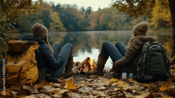 Fototapeta Two friends sitting by a cozy campfire near a lake, surrounded by autumn leaves, enjoying nature.