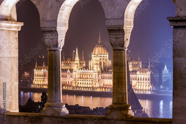 Fototapeta Night view of the Hungarian Parliament Building between the columns of the fortress and viewpoint of the Fisherman's Bastion, located on the shore of Buda
