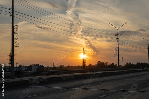 Fototapeta Orange sunset against of a power transmission tower. Cloudy sky. Asphalt road, pillars, horizon in the industrial part of the city. The Far North of Russia.